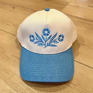 May include: A baseball cap with a white crown and a blue brim. The cap features a blue floral embroidery design on the front. The cap has a blue button on top.