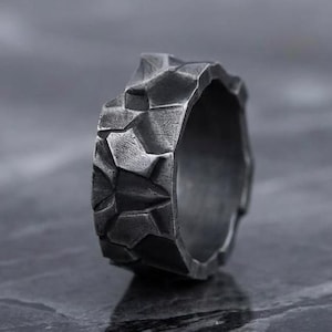 May include: A close-up of a textured, dark gray ring. The ring has a rugged, stone-like appearance with irregular facets. The ring is made of a dark metal and sits on a gray surface.