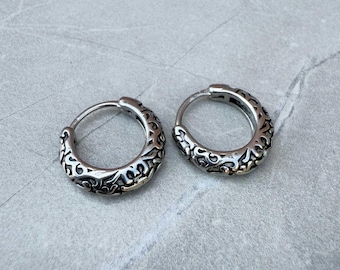 May include: A pair of silver hoop earrings with an intricate, ornate design. The earrings are made of metal and have a polished finish.