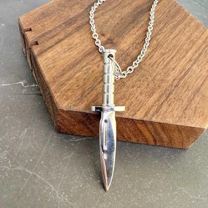 Puede incluir: Un collar con un colgante de cuchillo de plata con una cadena. El cuchillo está detallado con una punta afilada y un mango texturizado.