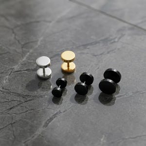 May include: An assortment of stud earrings in silver, gold, and black. The earrings have a dumbbell shape with a flat disc on each end, connected by a short post. The earrings are displayed on a gray surface.