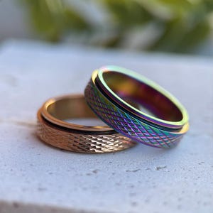 Peut inclure: Deux bagues aux surfaces texturées. L'une est dorée avec une bande noire, l'autre présente une finition irisée arc-en-ciel. Les bagues sont proches l'une de l'autre, mettant en valeur leurs motifs complexes et leur éclat métallique.
