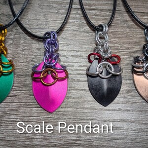 May include: Four scale pendants on black cords. The pendants have a shield shape with various colors: green, pink, black, and copper. The pendants are connected to the cord with metal chainmail. The text "Scale Pendant" is at the bottom.