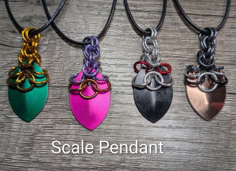 May include: Four scale pendants on black cords. The pendants have a shield shape with various colors: green, pink, black, and copper. The pendants are connected to the cord with metal chainmail. The text "Scale Pendant" is at the bottom.