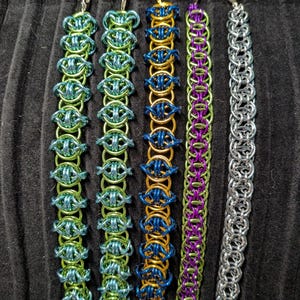 May include: Five chainmaille bracelets with lobster claw clasps. From left to right: a 6-inch teal and light green bracelet, a 7.25-inch light green and teal bracelet, a 9-inch gold and blue bracelet, an 8-inch purple and green bracelet, and an 8-inch silver bracelet.