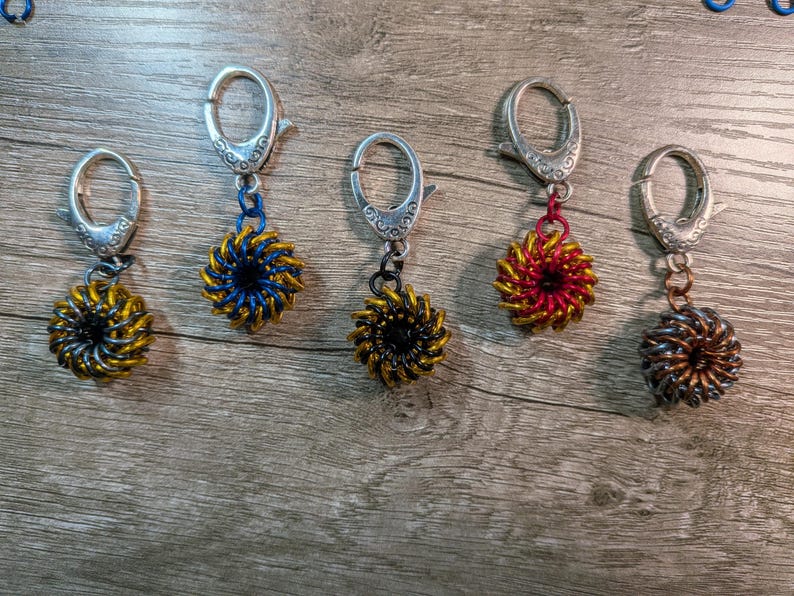 May include: Five handmade keychains with silver-colored clasps and spiral-shaped pendants. The pendants are in various colors, including gold, blue, black, red, and bronze. The keychains are displayed on a wooden surface.