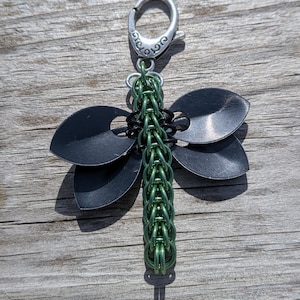 May include: A dragonfly-shaped keychain with a silver clasp. The wings are black, and the body is green. The word "Dragonfly" is written in white at the bottom.
