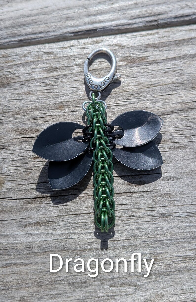 May include: A dragonfly-shaped keychain with a silver clasp. The wings are black, and the body is green. The word "Dragonfly" is written in white at the bottom.