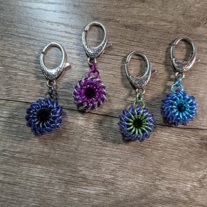 May include: Four handmade keychains with decorative floral-shaped pendants. The pendants are made of woven metal rings in shades of blue, purple, and green. Each keychain has a silver-colored clasp.