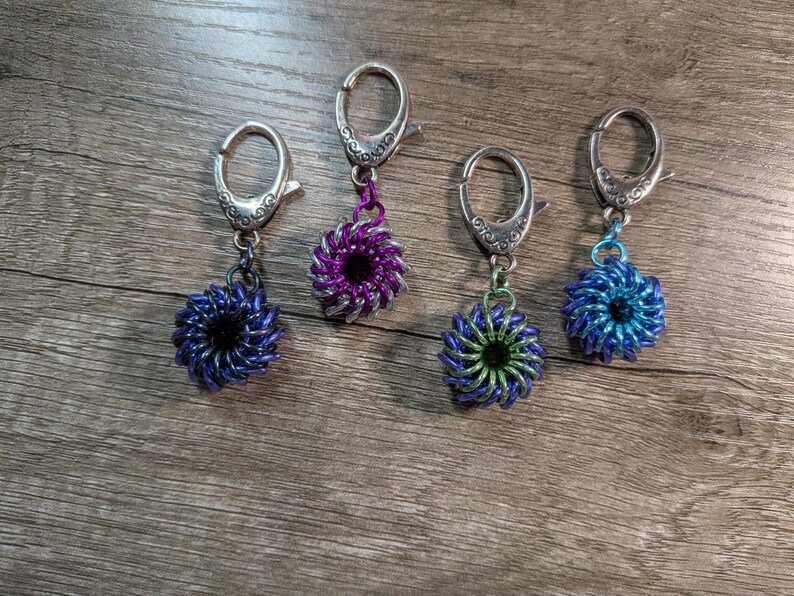 May include: Four handmade keychains with decorative floral-shaped pendants. The pendants are made of woven metal rings in shades of blue, purple, and green. Each keychain has a silver-colored clasp.