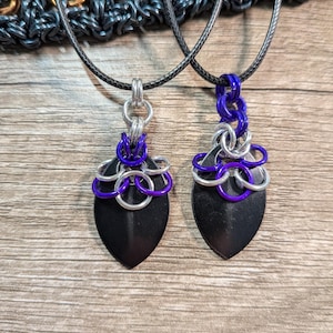 May include: Two pendant necklaces with black, shield-shaped pendants. Each pendant is adorned with silver and purple chainmail accents and hangs from a black cord. The background is a wooden surface.