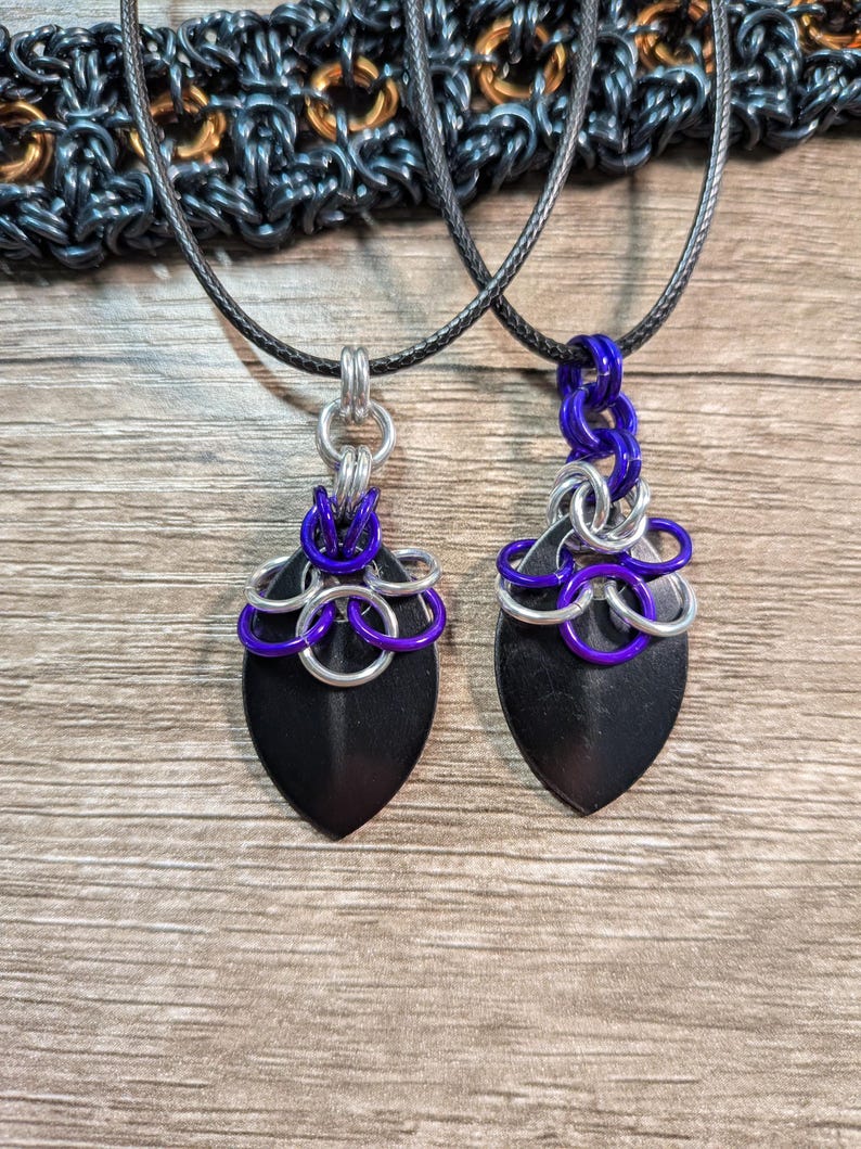 May include: Two pendant necklaces with black, shield-shaped pendants. Each pendant is adorned with silver and purple chainmail accents and hangs from a black cord. The background is a wooden surface.