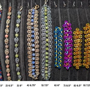 May include: Nine chainmaille bracelets arranged vertically. 1) Red, blue, silver 6.5 inches. 2) Pink, silver 6.5 inches. 3) Green, silver 8 inches. 4) Blue, gold 6.75 inches. 5) Green, silver 7.5 inches. 6) Blue, yellow 6 inches. 7) Purple, orange 8.5 inches. 8) Gold, green 6.5 inches. 9) Gold, green 7.25 inches.