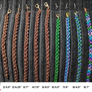 May include: Nine chainmaille bracelets with lobster claw clasps, arranged vertically. Five are copper-colored, one is green, one is blue and green, one is blue and purple, and one is a multi-colored gradient. Lengths range from 5.5 to 8.5 inches.