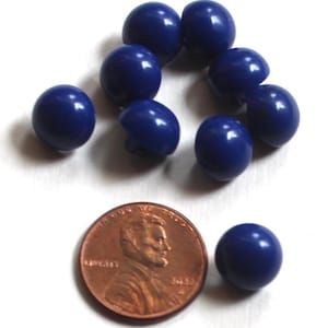May include: A collection of navy blue, round buttons. The buttons are arranged in a circular pattern, with one button and a penny coin placed nearby for size comparison. The buttons have a glossy finish and a classic design.