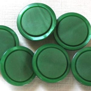 May include: A collection of six circular green buttons, each featuring a smooth, flat center surface surrounded by a slightly raised, rounded outer rim. The buttons are uniform in color and shape, displaying a consistent matte finish throughout.