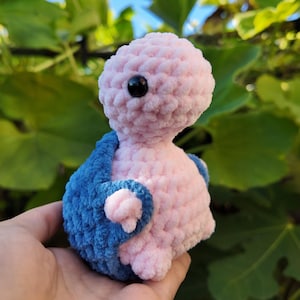 May include: A pink and blue crocheted stuffed animal with a single black eye. The animal is wearing a blue crocheted jacket.