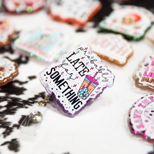 May include: A white and purple glitter badge holder with a clear plastic retractable reel. The badge holder features a cartoon image of a pink and orange iced coffee with the text "probably late for something".