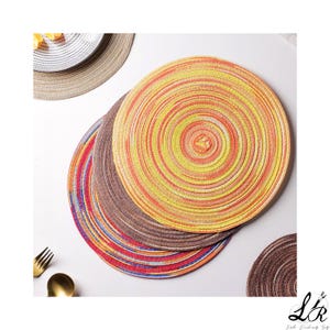 Op de afbeelding: Een verzameling kleurrijke ronde placemats. De placemats hebben concentrische cirkels in bruin-, rood-, geel- en oranje tinten. Een gouden vork en lepel zijn zichtbaar. De placemats zijn ontworpen voor de eetkamer.