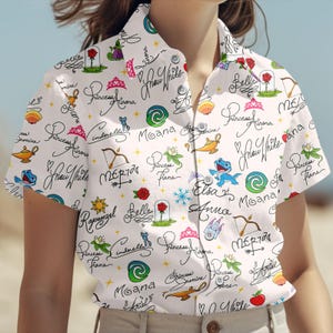 May include: White button-down shirt featuring a repeating pattern of Disney princess names and related illustrations. The design includes images of roses, tiaras, and characters like Elsa and Moana. The shirt has short sleeves.