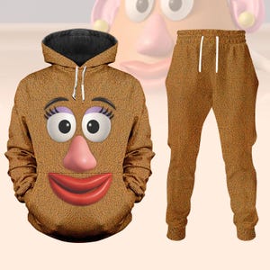 Può includere: Un completo marrone composto da felpa con cappuccio e pantaloni della tuta abbinati. La felpa con cappuccio presenta un volto di cartone animato con grandi occhi, un naso rosa e labbra rosse. La felpa ha un interno nero e coulisse bianche.