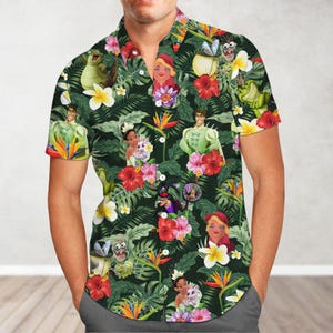 May include: A green short-sleeved button-down shirt with a tropical print featuring characters from the Disney movie The Princess and the Frog. The print includes Tiana, Naveen, Ray, Mama Odie, and Louis. The shirt has a green background with palm leaves, flowers, and other tropical elements.