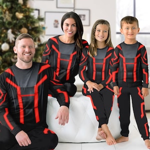 Puede incluir: Una familia de cuatro personas con pijamas negros a juego con diseños geométricos rojos. Los pijamas incluyen tops de manga larga y pantalones. La familia está sentada en un otomán blanco en una habitación bien iluminada.
