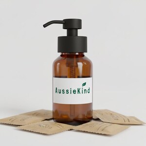 May include: An amber-colored glass bottle with a matte black pump dispenser. A white rectangular label on the front features the green text AussieKind with a small green leaf icon above the letter i. Several small brown paper packets surround it.