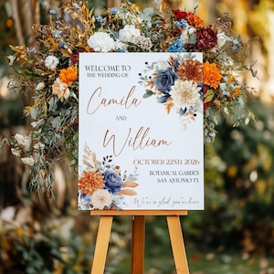 Op de afbeelding: Een wit trouwbord op een houten ezel, met bloemstukken in blauw, oranje en crème. Het bord leest: "Welkom bij de bruiloft van Camila en William, 22 oktober 2026, Botanische tuin, San Antonio TX."