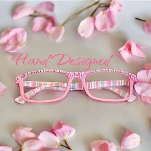 May include: Pink eyeglasses with a patterned design and rhinestones on the frame. The text "Hand Designed" is written above the glasses.