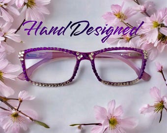 May include: Purple eyeglasses with rhinestones around the frame. The glasses are on a white background with pink flowers.