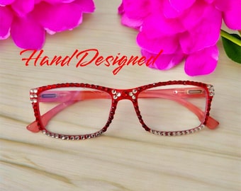 May include: Red eyeglasses with clear lenses and rhinestones around the frame. The text "Hand Designed" is visible above the glasses.
