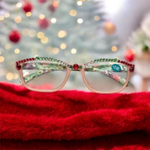May include: Reading glasses with a light pink frame, adorned with red and green gemstones along the top rim. The temples feature a floral pattern. The lenses have a +2.00 strength sticker. The glasses rest on a red surface.