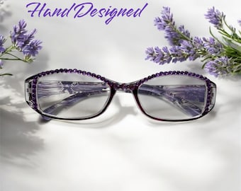 May include: Purple glasses with rhinestones around the frame. The glasses are on a white surface with lavender flowers.
