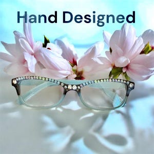 May include: A pair of eyeglasses with a light blue frame and embellished with rhinestones along the top edge. The text "Hand Designed" is at the top. Pink and white flowers are in the background.