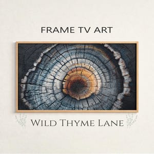 May include: Framed TV art print featuring a close-up of a tree cross-section. The artwork displays concentric rings in shades of gray, blue, and brown, with the text "FRAME TV ART" and "WILD THYME LANE". The frame is a light brown color.