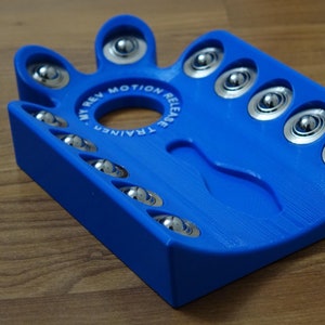 May include: Blue plastic device with a circular track and multiple ball bearing slots. The device is labeled "My Rev Motion Release Trainer".