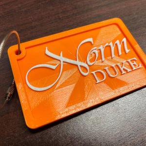 Puede incluir: Etiqueta de equipaje naranja con el nombre "Form Duke" en blanco, con un lazo de alambre plateado adjunto. La etiqueta tiene una superficie texturizada y un diseño geométrico.