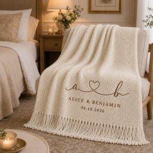 May include: A cream-colored knit throw blanket featuring a fringe edge and brown embroidered text that reads "a [heart symbol] b", "ALICE & BENJAMIN", and "06.18.2026". The blanket has a textured, chunky knit pattern and is draped over a light-colored chair.
