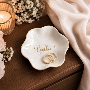 May include: A white, scalloped-edge ceramic trinket dish with a marble-like finish and a gold-painted rim. The centre features the name Cynthia in gold script lettering, flanked by two small gold hearts. Two gold rings rest inside the dish.