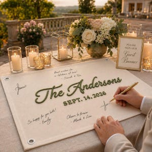 May include: A rectangular cream-colored fabric guest banner with metal grommets in the corners. It features the embroidered green text The Andersons and SEPT. 14, 2026, alongside handwritten signatures and messages in black ink on the surface.