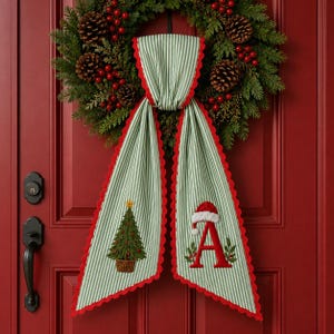 May include: A green and white striped fabric door hanger with a scalloped red edge. One side features an embroidered Christmas tree in a basket, while the other side displays a large red letter A topped with a miniature white-trimmed Santa hat.
