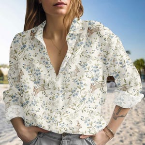 Puede incluir: Una camisa de manga larga con botones con estampado floral y de conejos. La camisa presenta un fondo color crema con flores azules y conejos marrones. Las mangas están enrolladas y la camisa tiene cuello en V.