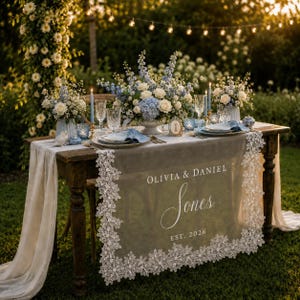 May include: A wooden table with a sheer white lace runner featuring the text OLIVIA & DANIEL Jones EST. 2026. The table is decorated with blue and white floral arrangements, blue taper candles, glassware, and blue napkins in an outdoor setting.