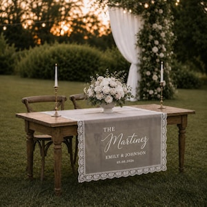 Peut inclure: Une table en bois avec un chemin de table en velours gris orné d'une bordure en dentelle blanche et du texte imprimé THE Martinez EMILY & JOHNSON 05.08.2026. La table porte un centre de table floral blanc et deux hauts bougeoirs en laiton.