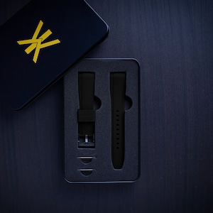 May include: A black watch strap set in a black box with a gold logo. The box is open, revealing two black watch straps and small metal pins. The background is a dark wood surface.