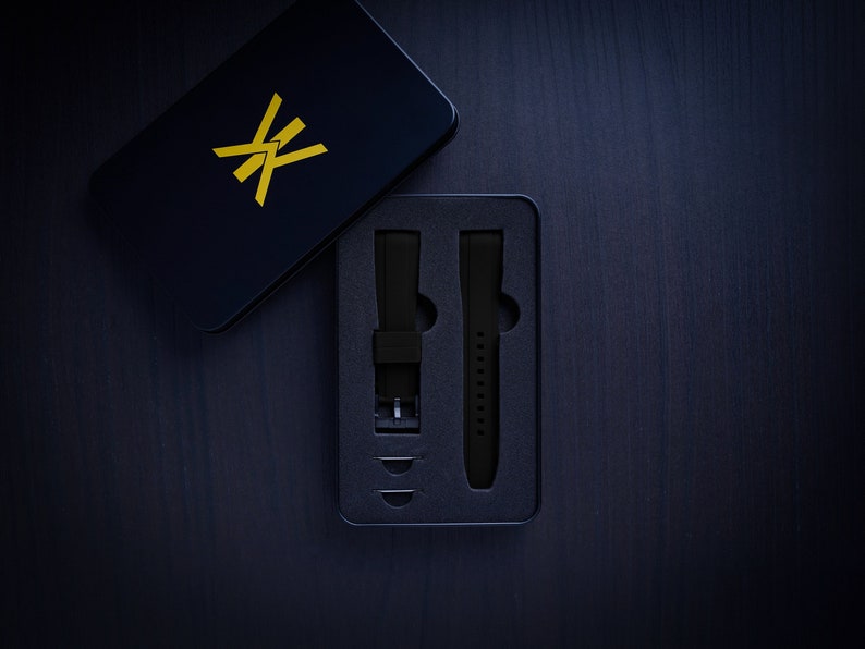 May include: A black watch strap set in a black box with a gold logo. The box is open, revealing two black watch straps and small metal pins. The background is a dark wood surface.