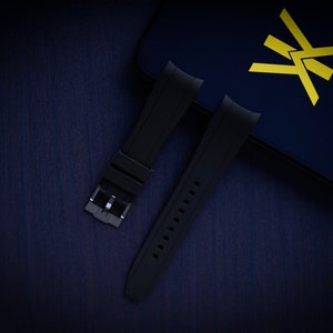 May include: Black watch straps with a silver buckle are displayed on a dark blue surface. The straps are made of a flexible material and have a series of holes for adjustment. A dark blue box with a yellow logo is in the background.