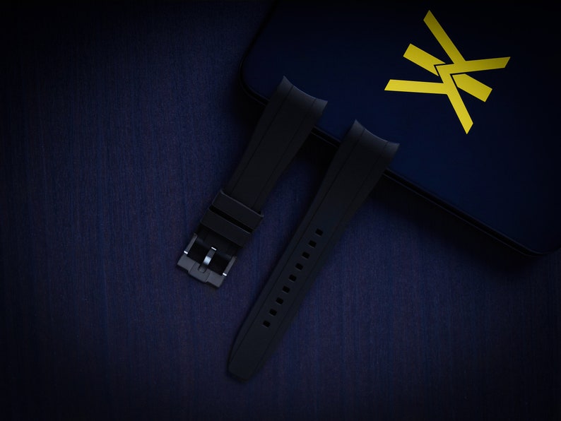 May include: Black watch straps with a silver buckle are displayed on a dark blue surface. The straps are made of a flexible material and have a series of holes for adjustment. A dark blue box with a yellow logo is in the background.