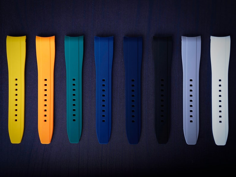 May include: A collection of seven watch straps in various colors, including yellow, orange, teal, blue, black, gray, and white. Each strap has a series of holes for adjustment and a curved design for the watch face.